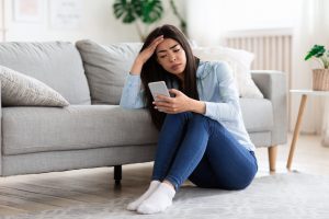 Smartphone Addiction And Depression. Upset Asian Girl Sitting On Floor At Home With Smartphone, Looking Sadly To Device Screen